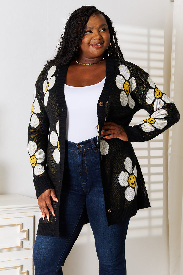 A person is smiling indoors against a white backdrop, wearing the Perfee Floral Button Down Longline Cardigan. It features large white flowers with yellow smiley faces on a black background. Their long hair complements the casual look, completed by a white top and blue jeans.