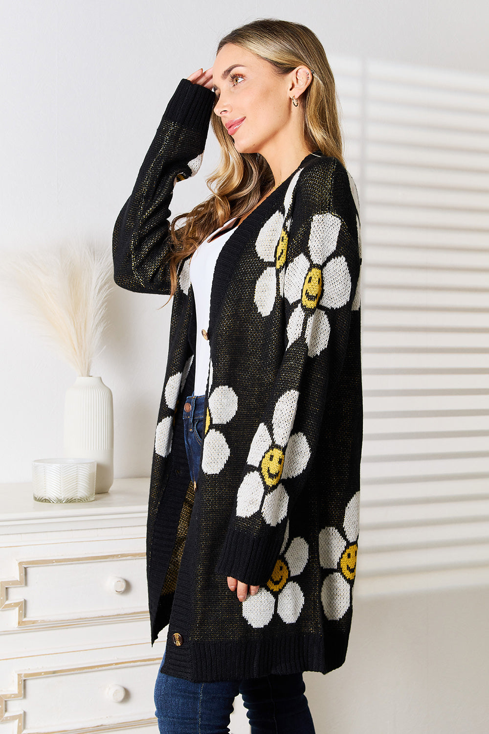 A woman stands in a room wearing the Perfee Floral Button Down Longline Cardigan, which showcases a pattern of large white smiling flowers. Her long, wavy hair cascades as she glances to the side. In the background, a white vase and a few drawers contribute to the serene setting.