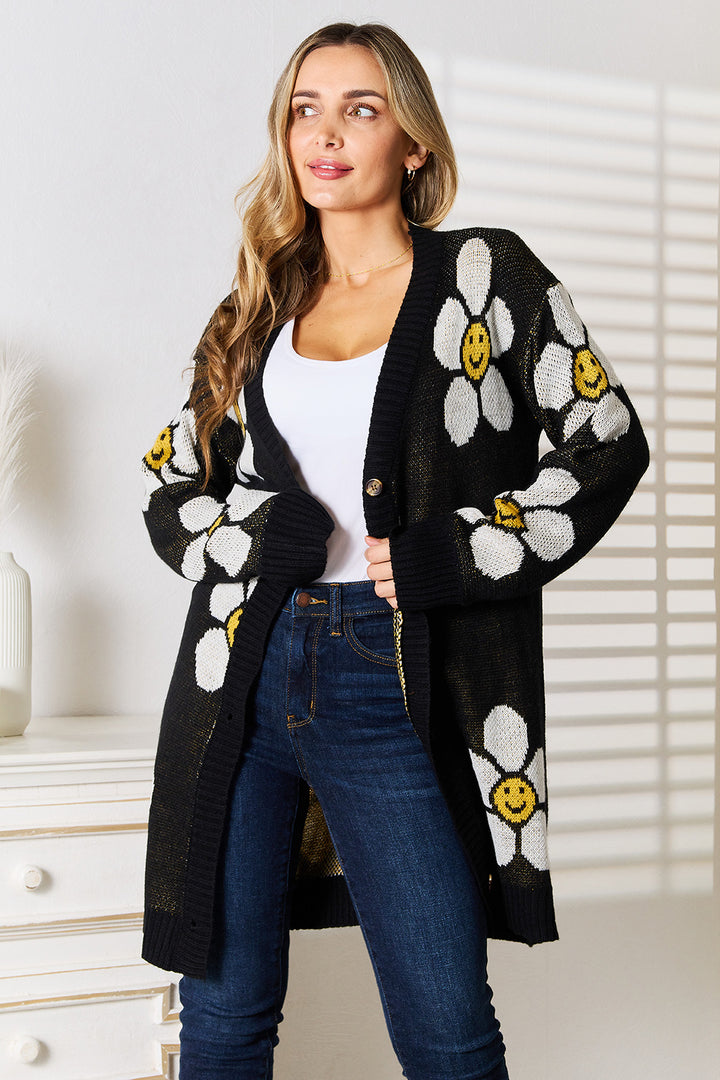 A woman stands in a room wearing the Perfee Floral Button Down Longline Cardigan, featuring a black base adorned with large white daisies and smiley faces. She has long hair and is dressed in a white button-down shirt and blue jeans, with a white cabinet holding a decorative vase visible in the background.