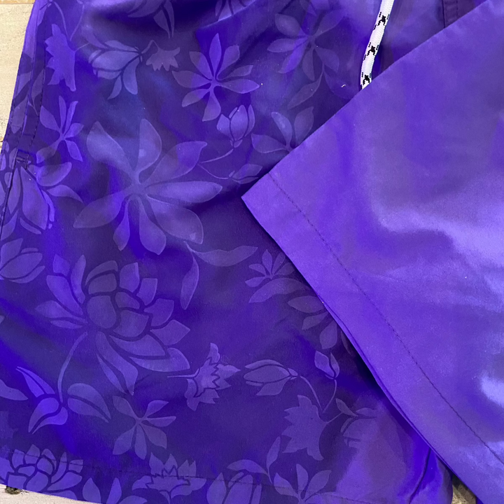 Close-up of Hibiscus Flower Pattern Changing Trunks Mens, featuring purple and blue overlapping hibiscus petals and leaves for a layered look. A contrasting white and black string is visible, adding a stylish touch to these swim trunks.