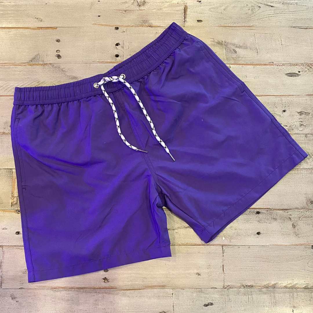 The Hibiscus Flower Pattern Changing Trunks for men, featuring purple fabric with a white and black drawstring, lie flat on a weathered wooden surface. These shorts with an elastic waistband exhibit a unique color-changing effect when wet.