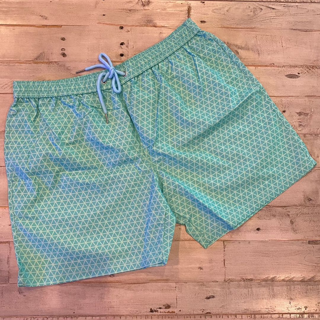 The Geo Pattern Men Color Changing Trunks, featuring a blue and green design with a drawstring, are laid flat on a wooden surface.