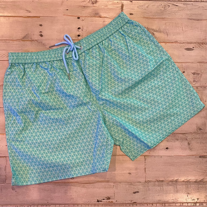 The Geo Pattern Men Color Changing Trunks, featuring a blue and green design with a drawstring, are laid flat on a wooden surface.