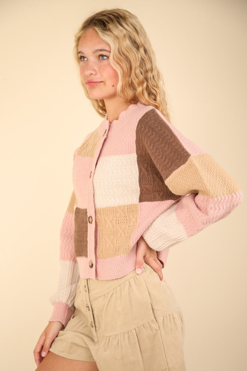 With long wavy hair, a person wears the VERY J Color Block Button Down Textured Sweater Cardigan, featuring pastel patchwork in shades of pink, brown, and beige. They pair it with light tan shorts against a plain beige background, creating a harmonious blend of subtle tones.