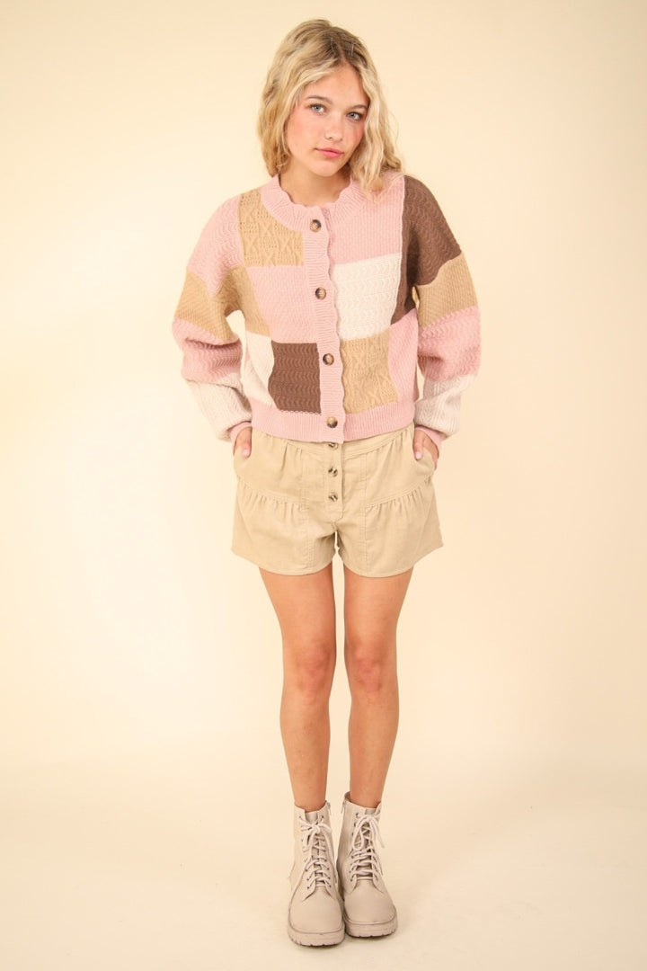 A person with short blonde hair is wearing the VERY J Color Block Button Down Textured Sweater Cardigan, along with beige shorts and beige boots. They stand against a plain light background, looking at the camera with hands in their pockets.