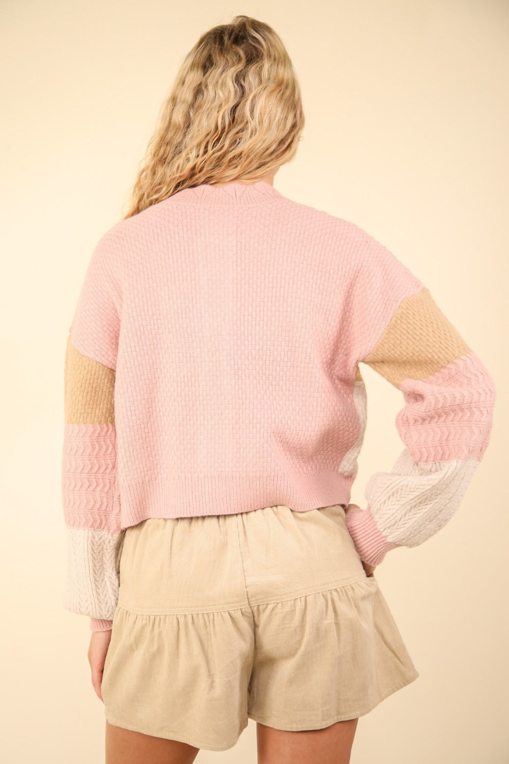 A woman with long blond hair stands with her back to the camera, wearing a VERY J Color Block Button Down Textured Sweater Cardigan in pink, beige, and cream, paired with beige shorts.