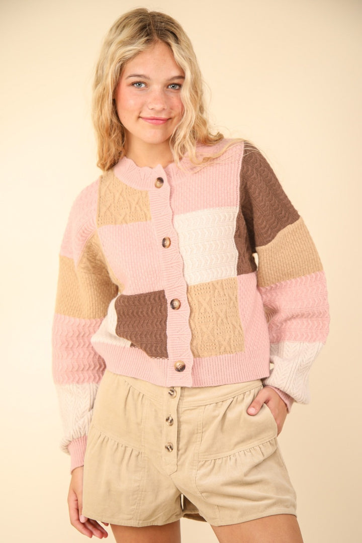 A person with blonde hair is wearing the VERY J Color Block Button Down Textured Sweater Cardigan, featuring shades of pink, brown, and cream with brown buttons, paired with beige shorts. The outfit stands out against a plain, light-colored background.