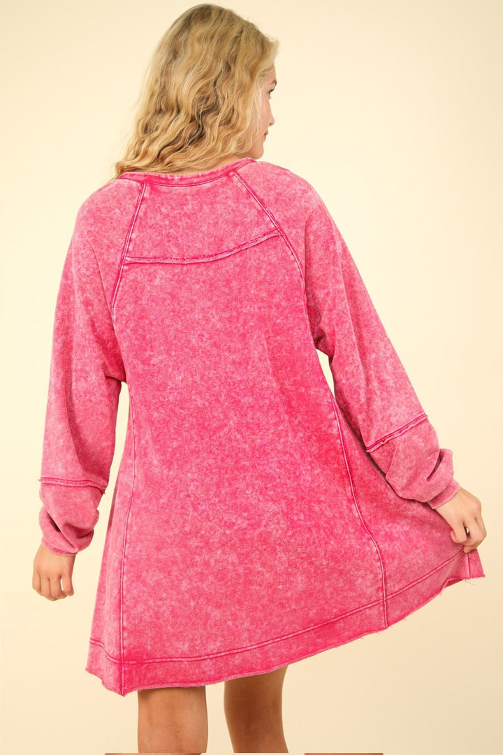 A person with long blonde hair wears the VERY J Mineral Washed Oversized A-Line Mini Dress in pink, featuring long sleeves. They stand back to the camera, gently holding one side of the dress, creating a vintage fashion vibe against a neutral background.