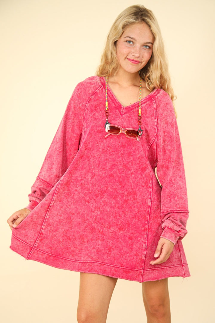 A person in a VERY J Mineral Washed Oversized A-Line Mini Dress with red long sleeves stands gracefully. The dress pairs with sunglasses hanging from a necklace, all against a simple, light background.