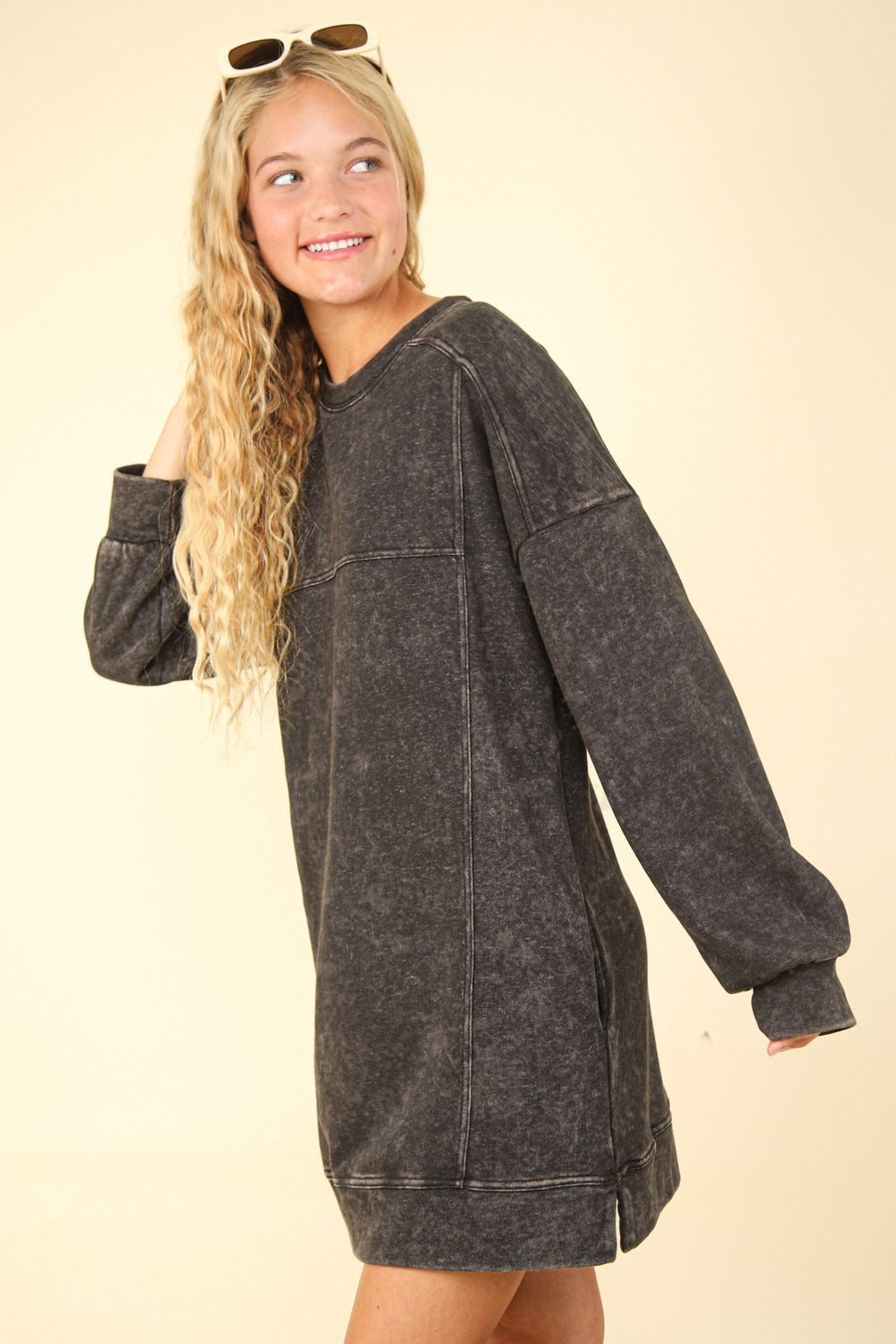 A person with long curly hair and sunglasses perched on their head smiles while looking to the side, dressed in a VERY J Mineral Washed Oversized Sweatshirt Mini Dress against a light background.