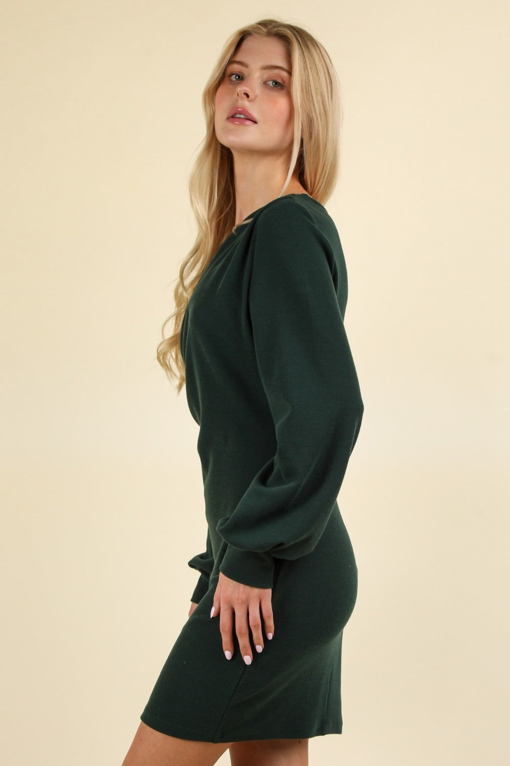 A woman with long blonde hair is wearing the VERY J Ruched Detail Bodycon Mini Dress, featuring a fitted silhouette, dark green color, long sleeves, and ruched detailing. She is standing sideways against a neutral background and looking forward.