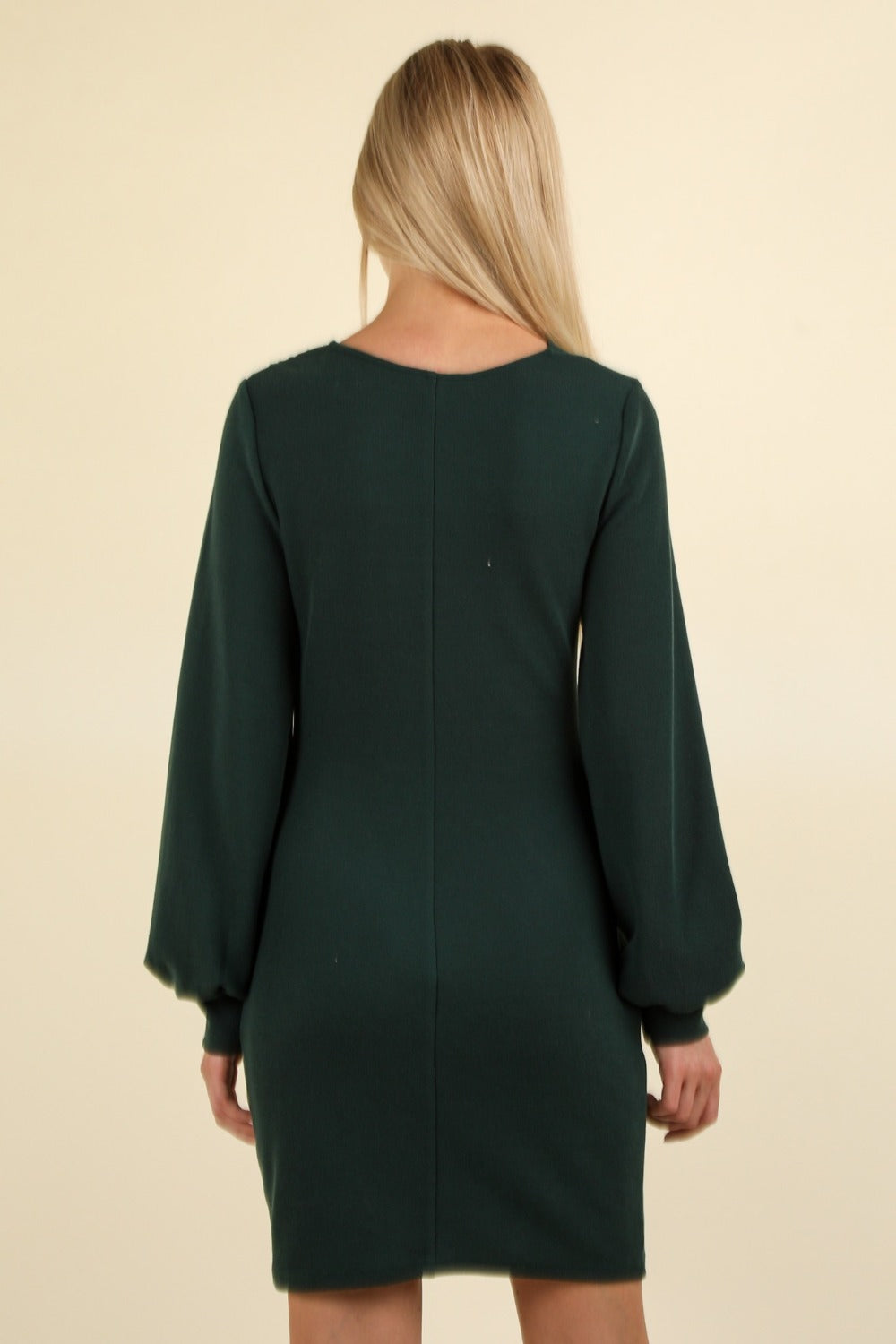 A person with long blonde hair stands with their back to the camera, wearing the VERY J Ruched Detail Bodycon Mini Dress in a dark green hue against a plain cream background.