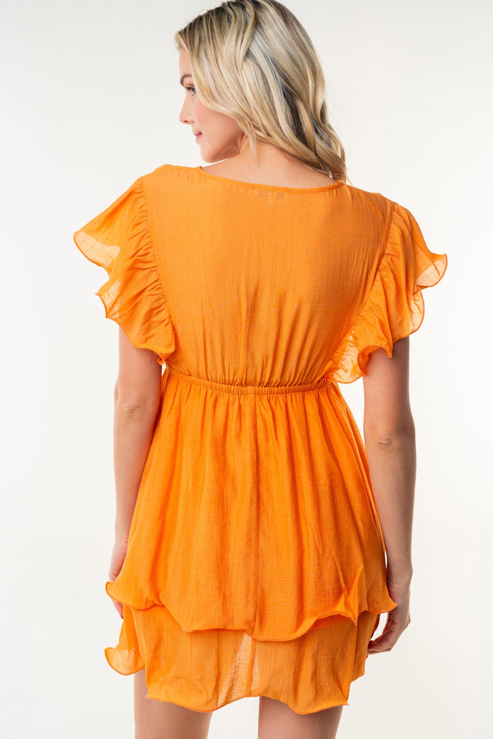 A woman with blonde hair is seen from behind, wearing the White Birch Full Size Short Sleeve Woven Layered Dress in orange, featuring ruffled sleeves and a tiered skirt. She stands against a plain white background.