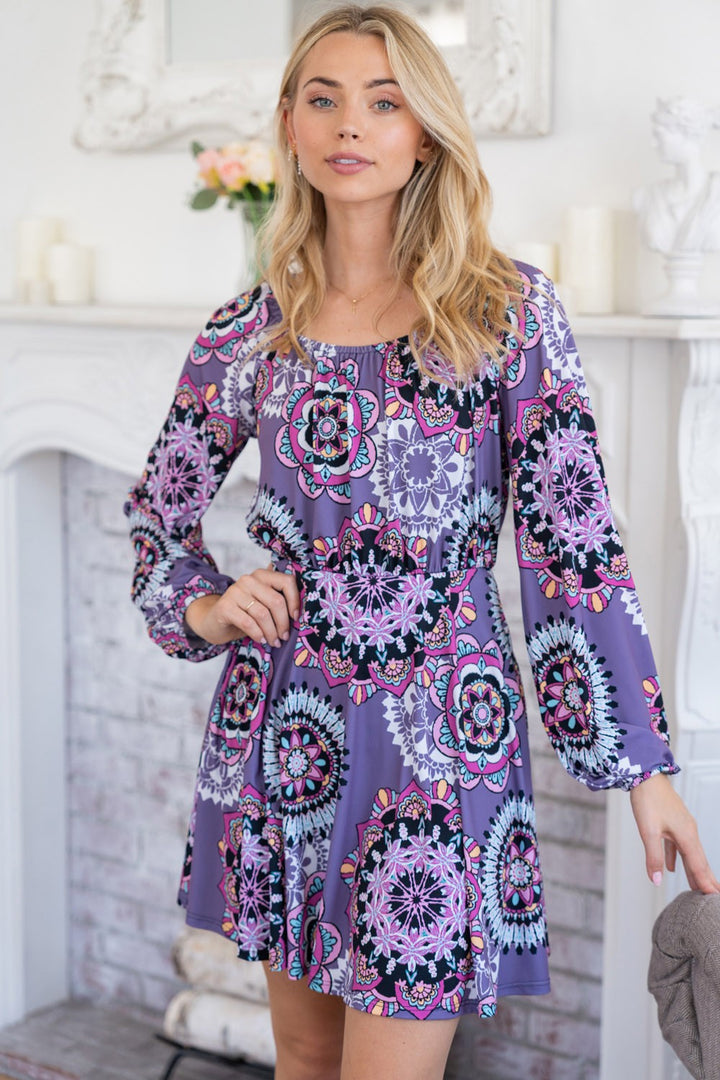 A woman poses indoors in a White Birch Printed Long Sleeve Mini Dress with Short Liner, featuring a vibrant mandala pattern and fitted waist. She stands with one hand on her hip in a room adorned with a white brick fireplace and decorative items.