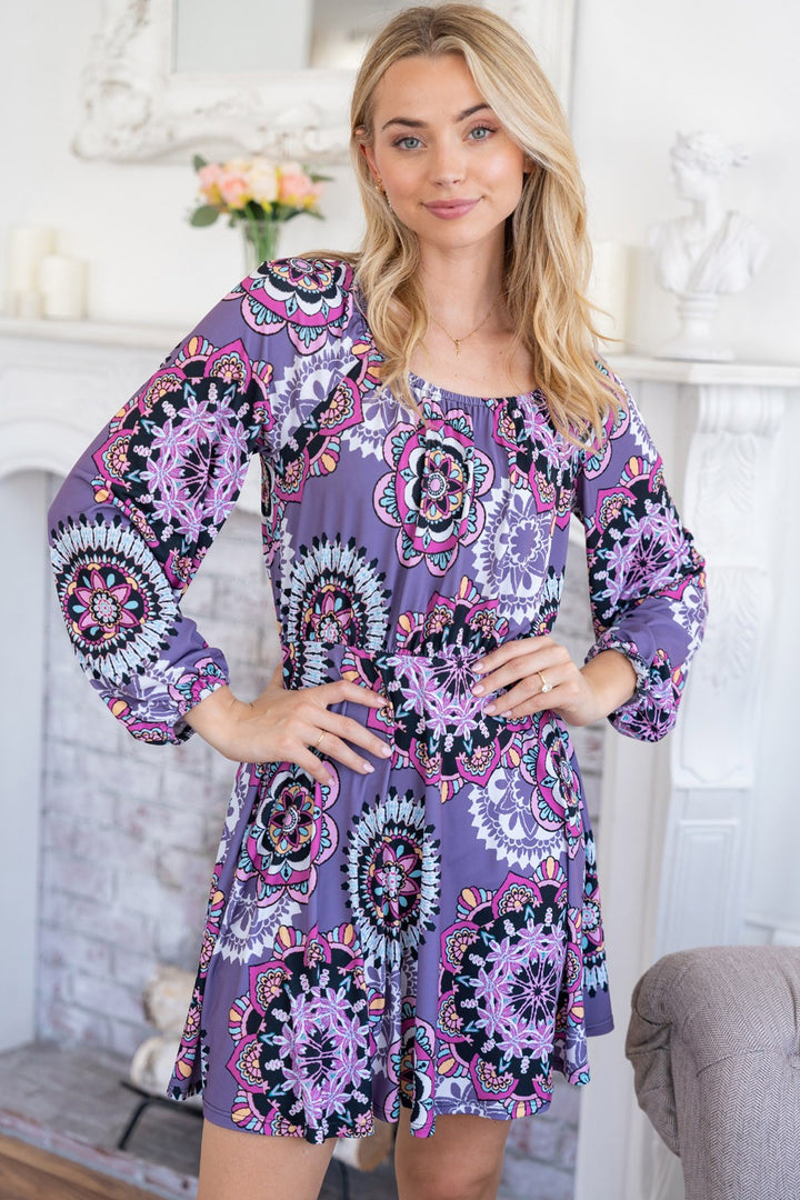 Indoors, a woman with blonde hair smiles, hands on hips, dressed in a White Birch Printed Long Sleeve Mini Dress with a vibrant purple floral pattern, standing in front of a decorative fireplace.