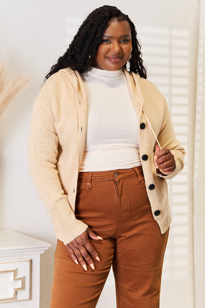 A person with long, curly hair is smiling while wearing a Woven Right Button-Down Long Sleeve Hooded Sweater over a white turtleneck and brown pants. They stand in a well-lit room with white walls and a decorative piece in the background, their cozy ensemble enhancing the serene atmosphere.