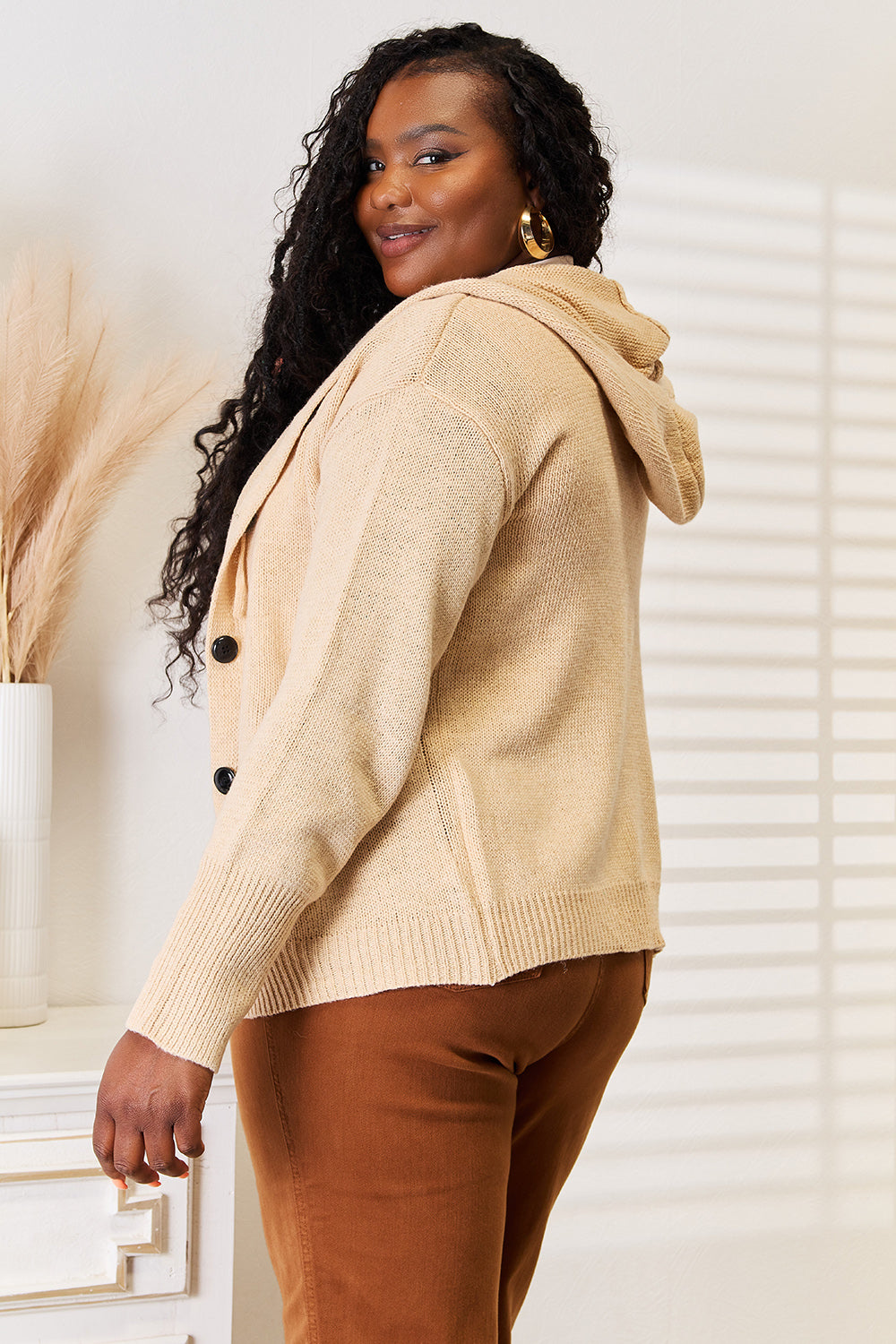 A person with long curly hair smiles while wearing the Woven Right Button-Down Long Sleeve Hooded Sweater in a tan shade, paired with brown pants, standing in a softly lit room adorned with dried pampas grass elegantly arranged in a vase.