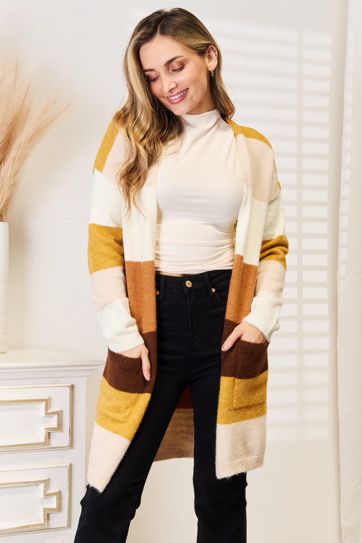 A woman stands smiling in a sunlit room, wearing the cozy Woven Right Color Block Dropped Shoulder Cardigan in shades of mustard, brown, and white over a white top. Her long, wavy blonde hair complements the black pants she sports as she stands next to a white dresser adorned with dried pampas grass.