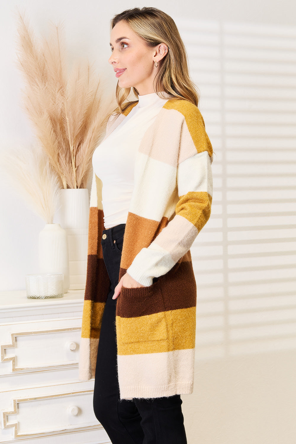 A person wearing the Woven Right Color Block Dropped Shoulder Cardigan in shades of white, brown, and mustard stands in profile. They have long hair and are dressed in a white top and black pants. The backdrop features dried pampas grass and a white chest of drawers.