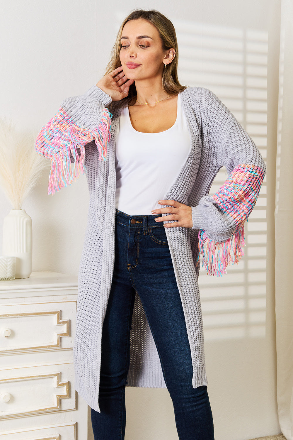 A woman poses indoors wearing the Woven Right Fringe Sleeve Dropped Shoulder Cardigan, a cozy, light gray knitted piece with vibrant fringed sleeves. It features a relaxed dropped shoulder design and pairs perfectly with her white top and dark jeans. A decorative vase in the background enhances the inviting ambiance.