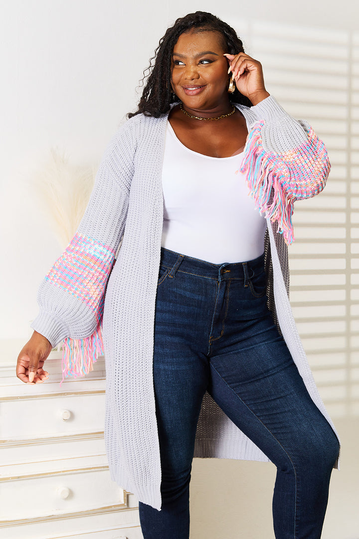 A person wearing a white top, blue jeans, and the Woven Right Fringe Sleeve Dropped Shoulder Cardigan with its colorful fringe sleeve detailing is smiling and posing in a brightly lit room.