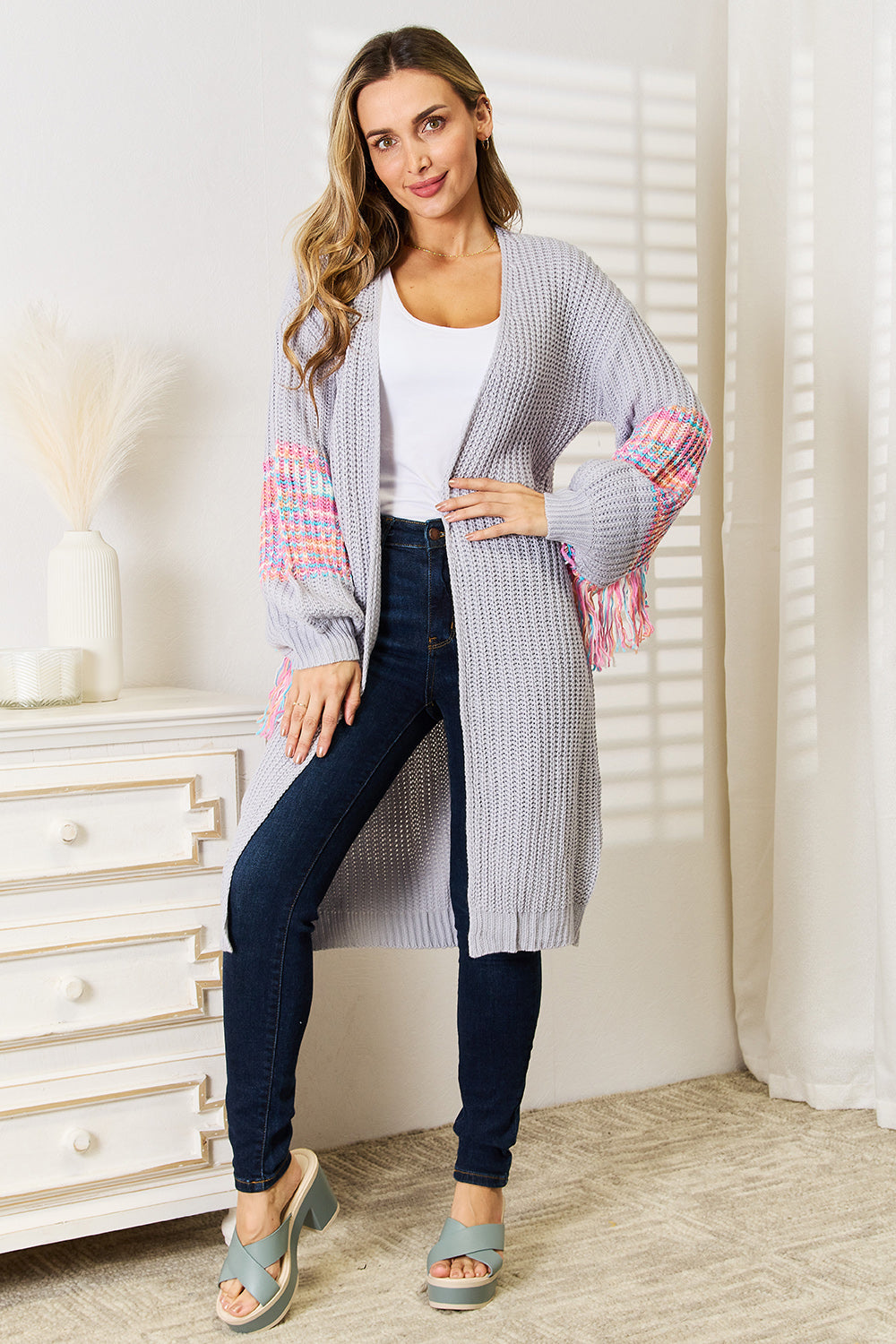 A woman stands by a white dresser, exuding style in her Woven Right Fringe Sleeve Dropped Shoulder Cardigan paired with a white top, dark jeans, and light green platform sandals. The cozy setting features minimalist decor that perfectly complements her chic ensemble.