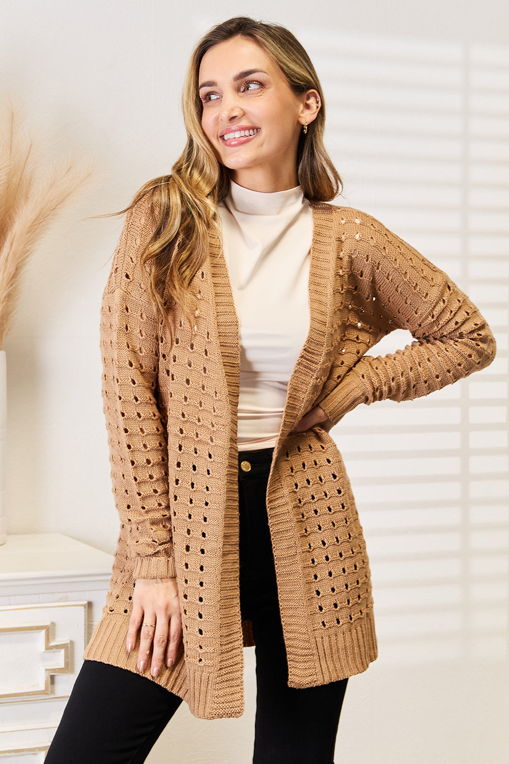 A person with long hair smiles while leaning against a textured wall, wearing the Woven Right Openwork Horizontal Ribbing Open Front Cardigan in beige over a white turtleneck and black pants. Soft lighting and shadows from window blinds create a cozy atmosphere.