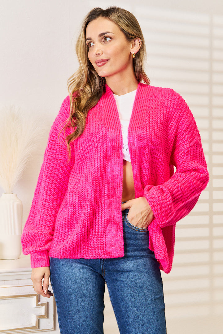 A person wearing the Woven Right Rib-Knit Open Front Drop Shoulder Cardigan in bright pink over a white top and blue jeans stands next to a white wall and a decorative vase with pampas grass. They have long hair and are gazing to the side with a relaxed expression.