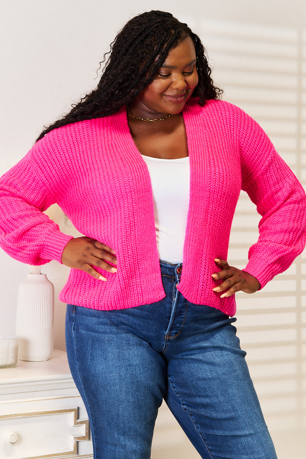 A person wearing the Woven Right Rib-Knit Open Front Drop Shoulder Cardigan in bright pink over a white top and blue jeans stands indoors, smiling with hands on hips. The background showcases light-colored walls and a shelf adorned with decorative items.