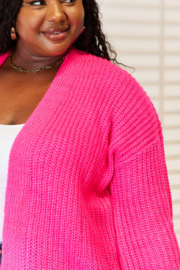 A person wearing a vibrant pink Woven Right Rib-Knit Open Front Drop Shoulder Cardigan smiles while looking to the side. They have long curly hair and small hoop earrings, and the background features softly blurred horizontal blinds.