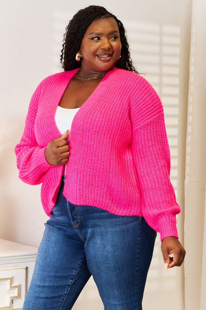 A person wearing the Woven Right Rib-Knit Open Front Drop Shoulder Cardigan in bright pink over a white top and blue jeans is smiling while looking to the side. They have curly hair and gold hoop earrings. The background is minimal and softly lit.