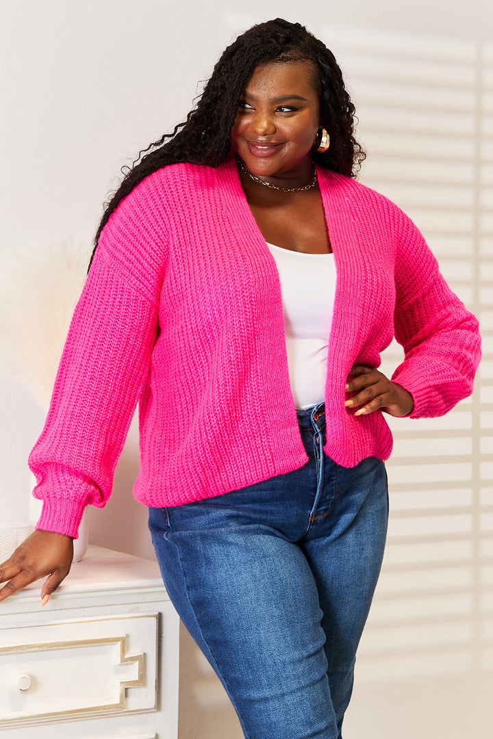 A person wearing the Woven Right Rib-Knit Open Front Drop Shoulder Cardigan in vibrant pink over a white top and blue jeans stands near a white cabinet. Their long, curly hair cascades beautifully as they smile while looking off to the side in the softly lit room, casting gentle shadows on the wall.