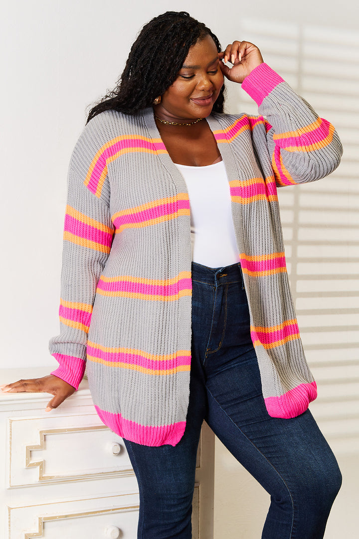 A person wearing a white top, blue jeans, and the Woven Right Ribbed Long Sleeve Cardigan featuring pink and orange stripes leans against a white dresser with a smile. This versatile wardrobe addition is complemented by the shadow pattern from window blinds in the background.