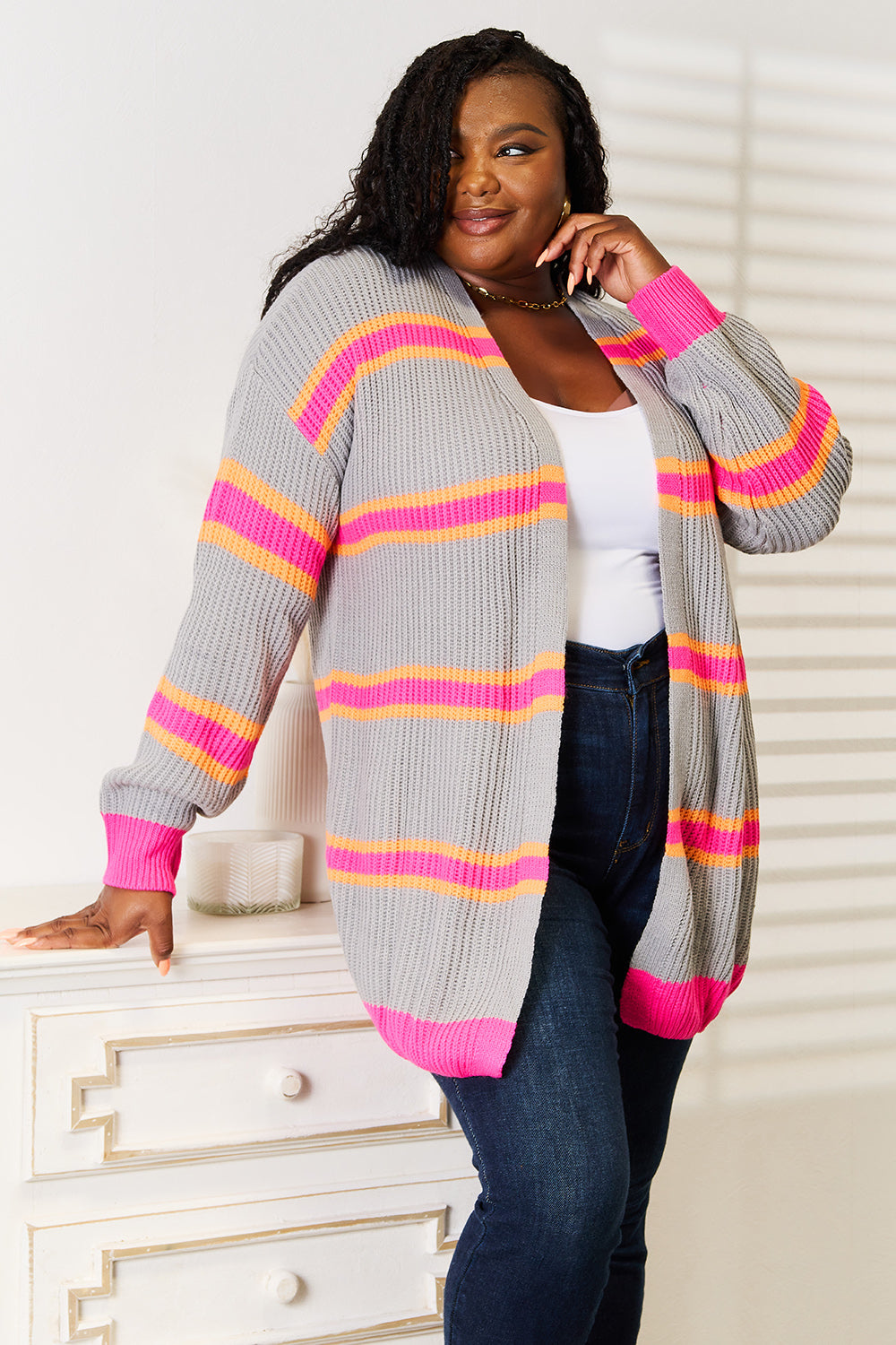 A person wearing a versatile wardrobe addition—a Woven Right Ribbed Long Sleeve Cardigan featuring gray with pink and orange stripes—paired with a white top and blue jeans, poses confidently. They lean against a white dresser, with sunlight casting shadows in the background.