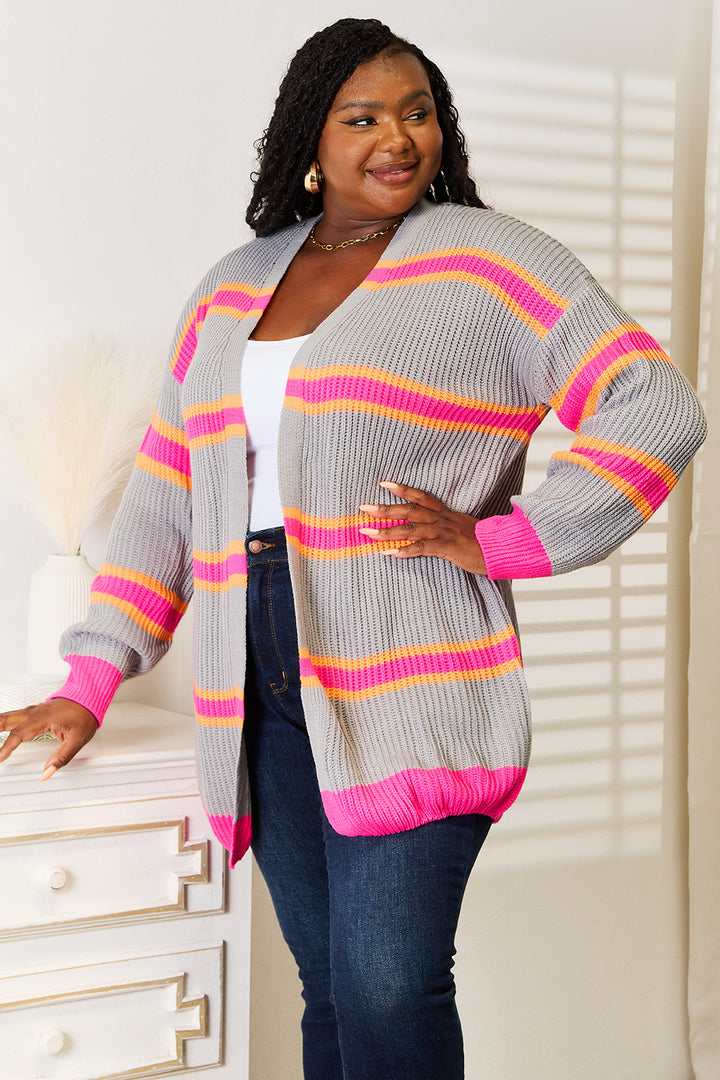 A person wearing the Woven Right Ribbed Long Sleeve Cardigan with pink and yellow stripes smiles while standing near a white dresser. They are also wearing a white top and blue jeans. A decorative vase with dried grass is on the dresser, showcasing how this versatile wardrobe addition complements cozy spaces.