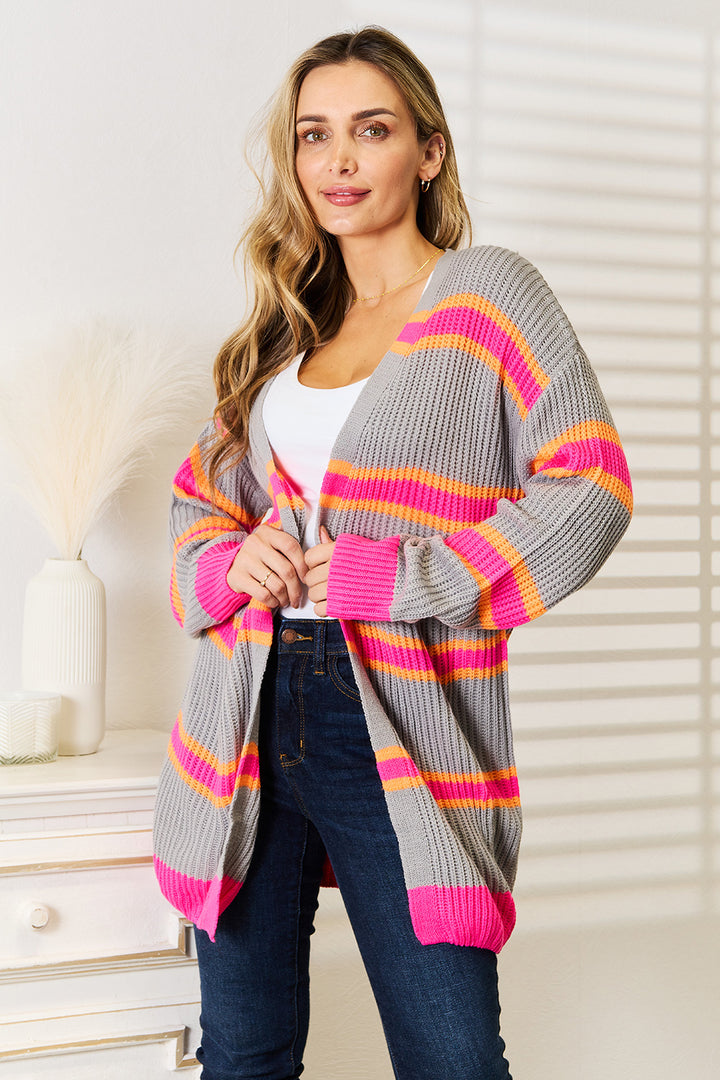 A person wearing the Woven Right Ribbed Long Sleeve Cardigan, a gray piece with pink and orange stripes, poses in a brightly lit room. They're standing near a white dresser adorned with decorative items, including a vase of fluffy white pampas grass. This versatile wardrobe addition effortlessly complements the serene setting.