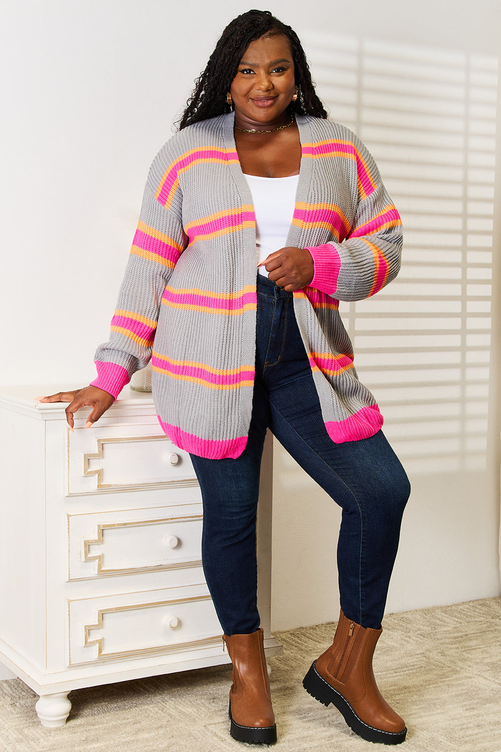 A woman stands casually indoors wearing the Woven Right Ribbed Long Sleeve Cardigan, featuring shades of grey, pink, and orange over a white top. She pairs it with dark jeans and brown boots. Leaning on a white dresser and smiling warmly, she showcases a versatile wardrobe addition.