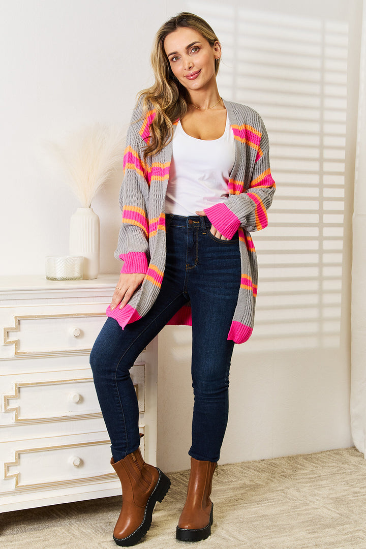 A person stands indoors, dressed in the Woven Right Ribbed Long Sleeve Cardigan featuring colorful stripes over a white top, paired with dark jeans and brown boots. They lean against a white chest of drawers adorned with a decorative vase, while sunlight filters through the blinds, casting delicate shadows on the wall.