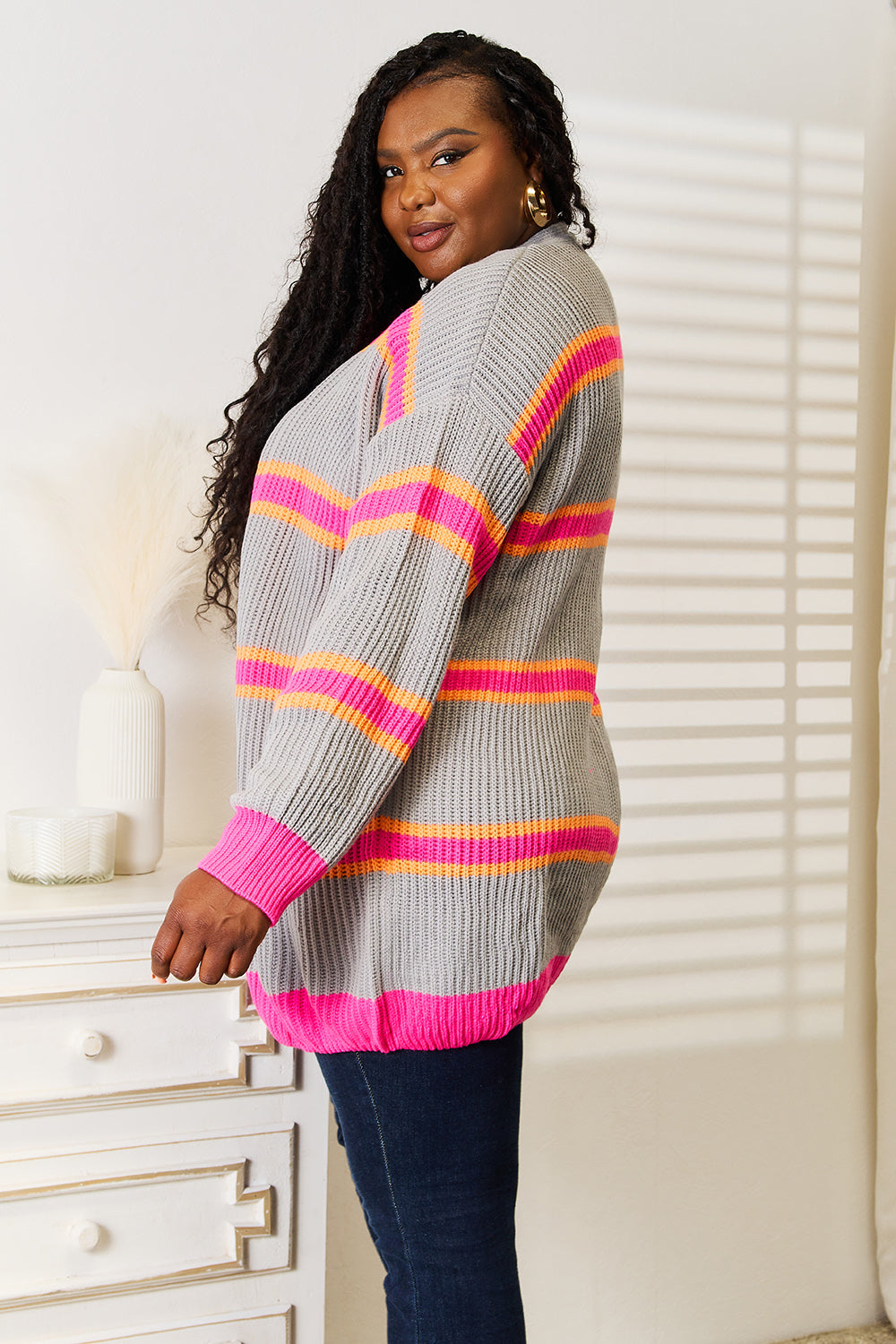 A person with long hair wearing the Woven Right Ribbed Long Sleeve Cardigan, featuring pink and orange stripes, stands near a white dresser. The sunlight casts striped shadows on the wall behind them, highlighting the cardigan as a versatile wardrobe addition.