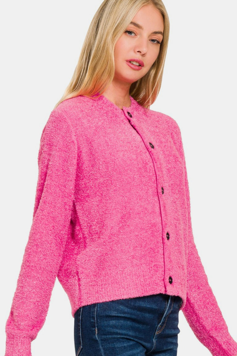 A woman with long blonde hair poses in the Zenana Button Down Long Sleeve Sweater Cardigan, its bright pink hue contrasting elegantly with her dark jeans, against a plain white background.