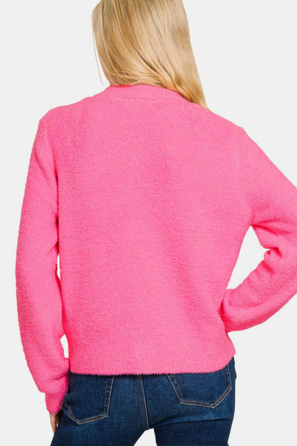 A person with long blonde hair is facing away, wearing a vibrant Zenana Button Down Long Sleeve Sweater Cardigan in pink over dark blue jeans. The background is plain white.