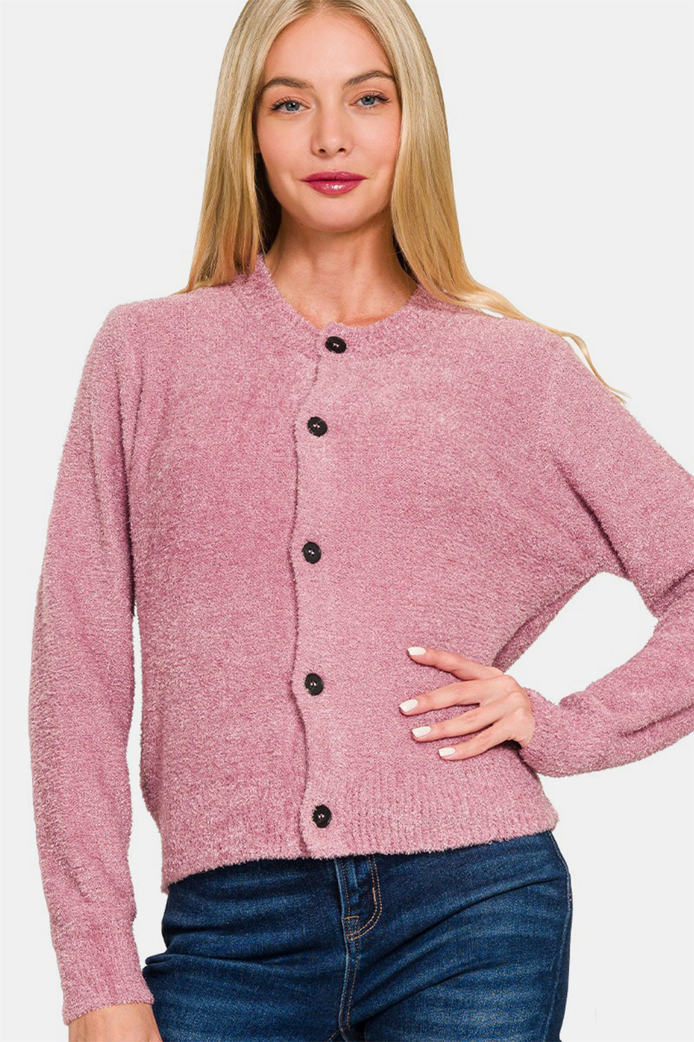 A person with long blonde hair is wearing the Zenana Button Down Long Sleeve Sweater Cardigan in pink, along with blue jeans, standing with one hand on their hip against a plain white background.