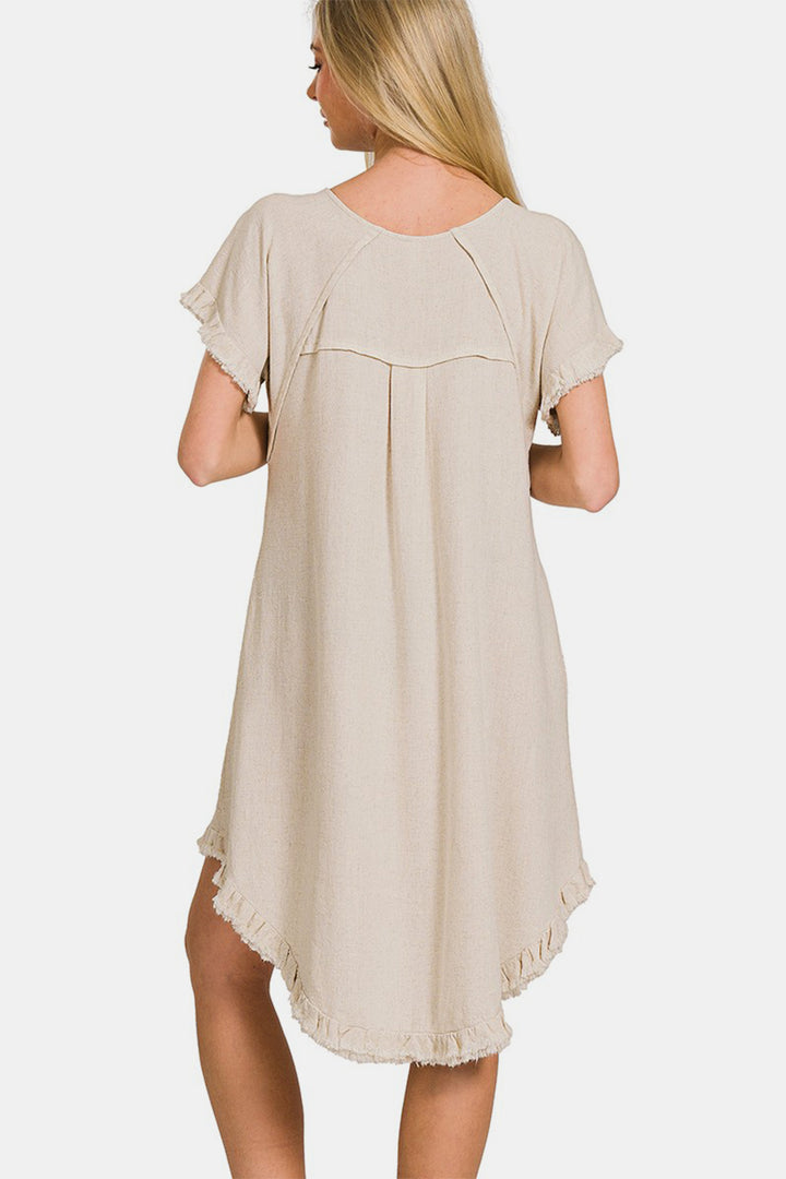 A woman with long blonde hair models the Zenana Fringe Edge High Low Flowy Dress with Pockets, featuring short sleeves and a frayed hem. She stands with her back to the camera against a white background.