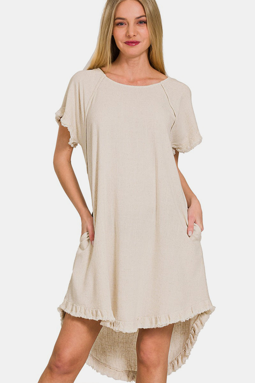 A woman in a Zenana Fringe Edge High Low Flowy Dress with pockets smiles with her hands in them. The beige dress has short sleeves, ruffled edges, and a high-low hem against a white background.