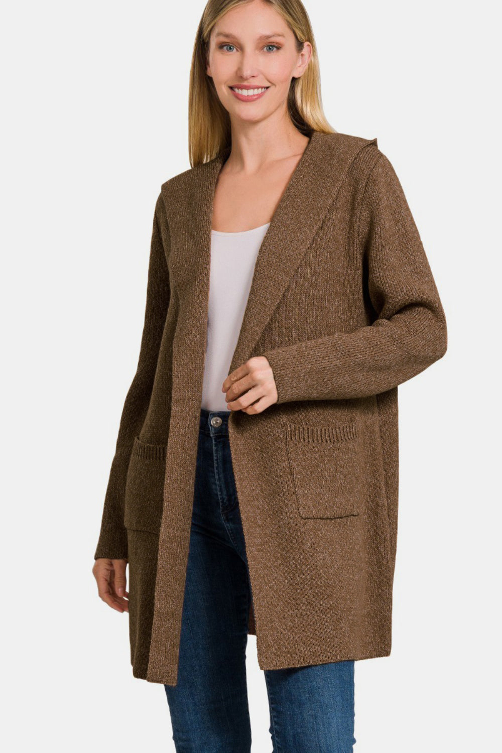 A person is standing against a plain background, smiling while wearing the Zenana Hooded Open Front Sweater Cardigan in brown over a white top and blue jeans. This cozy cardigan includes two front pockets and offers a relaxed fit.