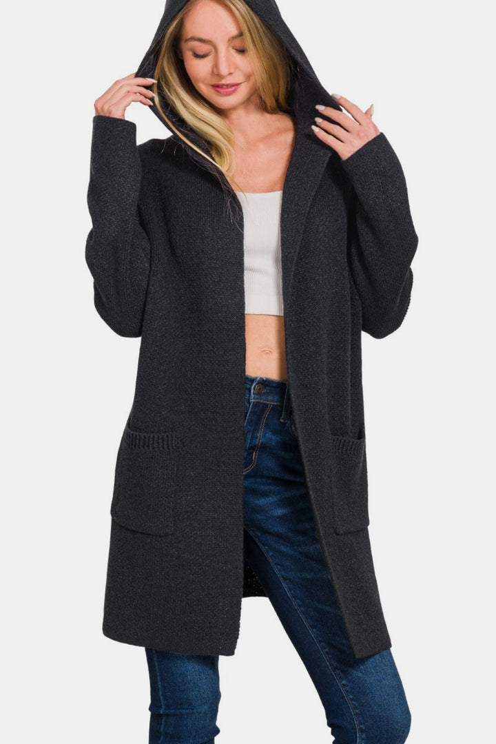 A person wearing a Zenana Hooded Open Front Sweater Cardigan in dark gray, ideal for cozy layering, stands with their hands adjusting the hood. The white top and blue jeans nicely complement the ensemble, and the front pockets add practicality. They are smiling gently.