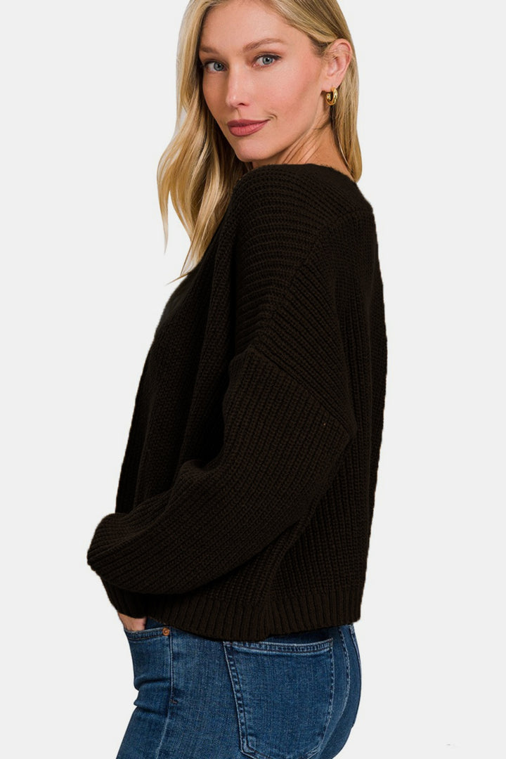 A woman wearing a Zenana Open Front Drop Shoulder Sweater Cardigan in black is layered over blue jeans, posing against a plain white background. She has long blonde hair, wears hoop earrings, and looks toward the camera with a slight smile.