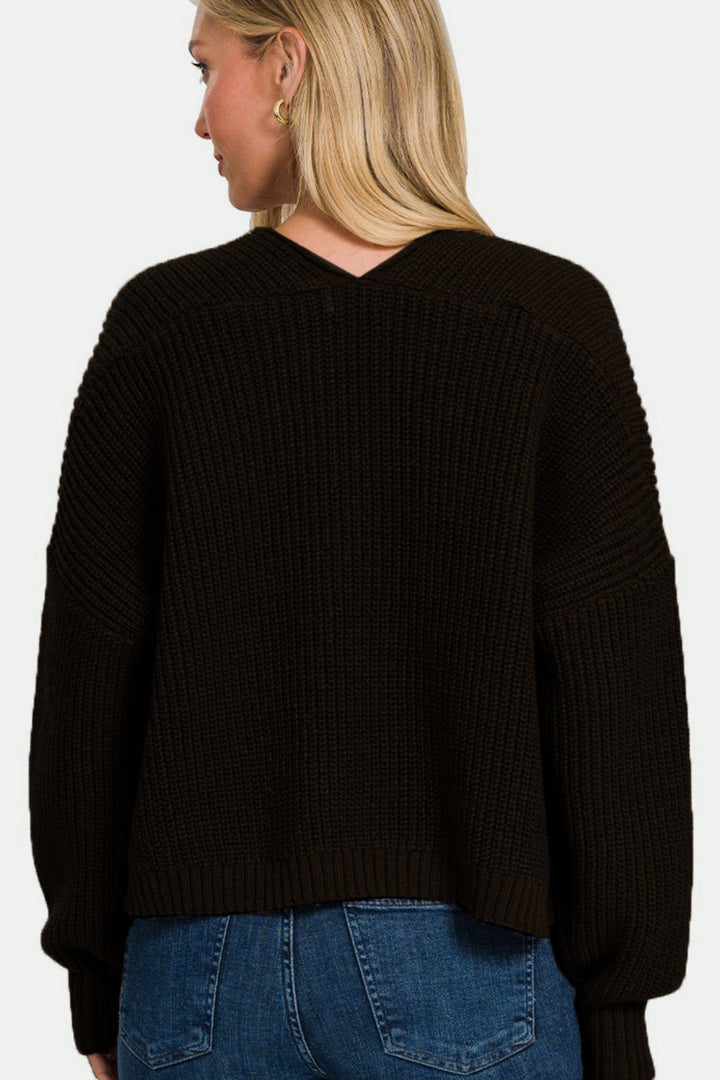 A person with long, blonde hair is wearing the Zenana Open Front Drop Shoulder Sweater Cardigan in black over denim jeans, facing away. The cardigan drapes elegantly with its textured knit and showcases a v-shaped neckline at the back.