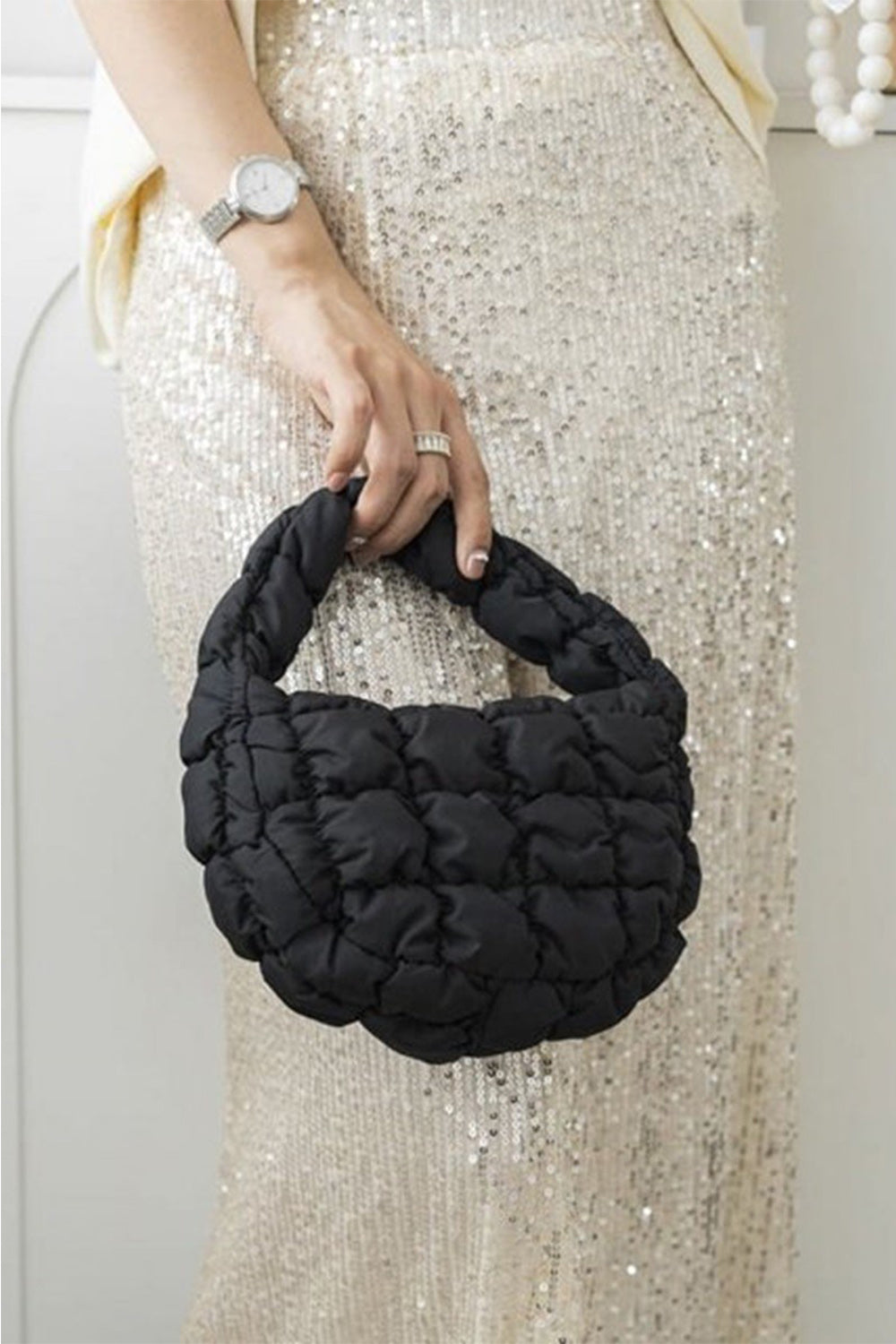 A person holds a Zenana Quilted Micro Puffy Handbag in their right hand, adding a stylish touch to their ensemble. They are wearing a sparkly, sequined skirt and a wristwatch. The setting appears indoors with a hint of a white wall in the background.