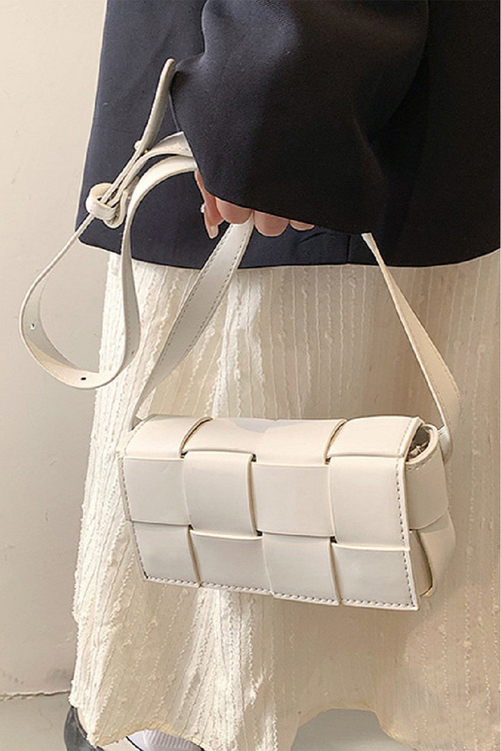 A person holds a Zenana Vegan Leather Woven Crossbody Bag with a long strap. Theyre dressed in a black jacket and textured, cream-colored skirt or dress against a neutral background.